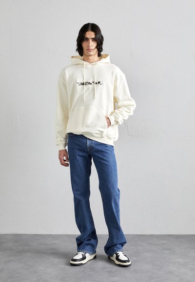 Cremefarvet hætte sweatshirt med frontlomme og sort tekstgrafik, parret med blå jeans og sort-hvide sneakers. Glat stof og afslappet pasform.