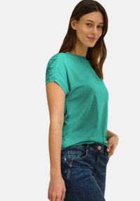 Camiseta de manga corta en color turquesa con detalle de fruncido en los hombros, combinada con jeans de mezclilla azul oscuro y un bolsillo en la parte frontal con botones.