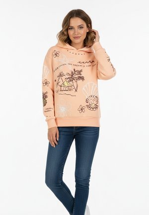 Woman wearing peach hoodie with tropical drawings, text "Everything you dream is coming" and blue jeans, standing against white background.