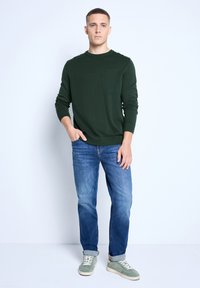 Un pull en laine vert foncé avec des poignets côtelés, associé à un jean bleu clair et des baskets vertes. Le modèle se tient de face avec une main dans la poche.