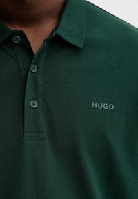 Polo de color verde oscuro hecho de algodón texturizado, con cuello, dos botones y un sutil logo "HUGO" en el lado izquierdo del pecho.