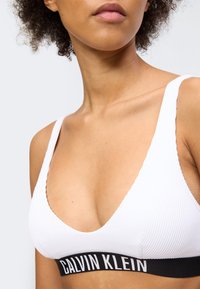 Naine, kes kannab valget soonikutega braletit musta Calvin Kleini elastse ribaga, näidates kaela, õlgu ja ülaosa rinnast.