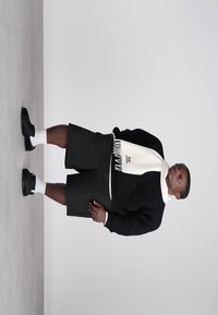 Mann iført svart genser, svarte shorts, svarte sko med hvite sokker, og et hvitt skjerf som står foran en enkel lys grå bakgrunn.