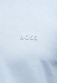 Λευκό υφασμάτινο ύφασμα με την λέξη «BOSS» να είναι ανάγλυφη σε λευκό, κεντραρισμένη στην επιφάνεια.