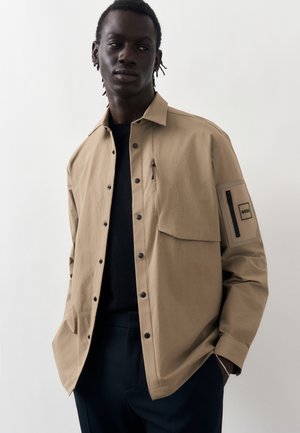 Homme portant une veste beige boutonnée avec une poche à rabat à l'avant et un patch zippé sur la manche, chemise noire et pantalon foncé sur fond uni.
