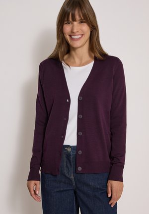 Femme souriante portant un cardigan boutonné violet foncé par-dessus une chemise blanche et un jean bleu, debout devant un fond clair uni.