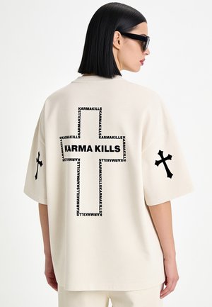 Frau mit schwarzen Haaren und Sonnenbrille trägt ein weißes oversized T-Shirt mit schwarzem Kreuz und "Karma Kills" Text auf dem Rücken sowie Kreuz-Designs auf den Ärmeln.