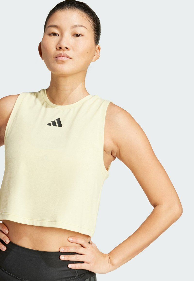 Top corto amarillo hecho de un tejido suave, con un logo negro de Adidas en el pecho. La modelo lleva leggings deportivos negros.