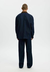 Personne debout de dos portant une veste en denim bleu foncé et un pantalon ample assorti sur un fond blanc uni.