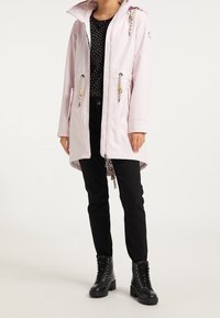 Veste imperméable rose clair avec fermeture éclair à l'avant, capuche ajustable et cordons décoratifs. Associée à un haut noir à pois et un pantalon noir.