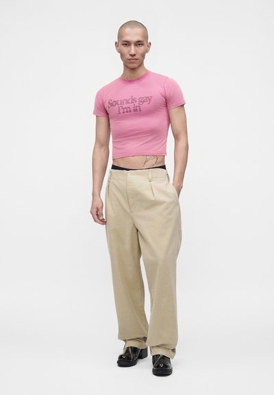 Νεαρό φαλακρό άτομο που φοράει ροζ κοντό t-shirt με κείμενο, μπεζ φαρδιά παντελόνια και μαύρα παπούτσια, στέκεται με το ένα χέρι στην τσέπη σε λευκό φόντο.