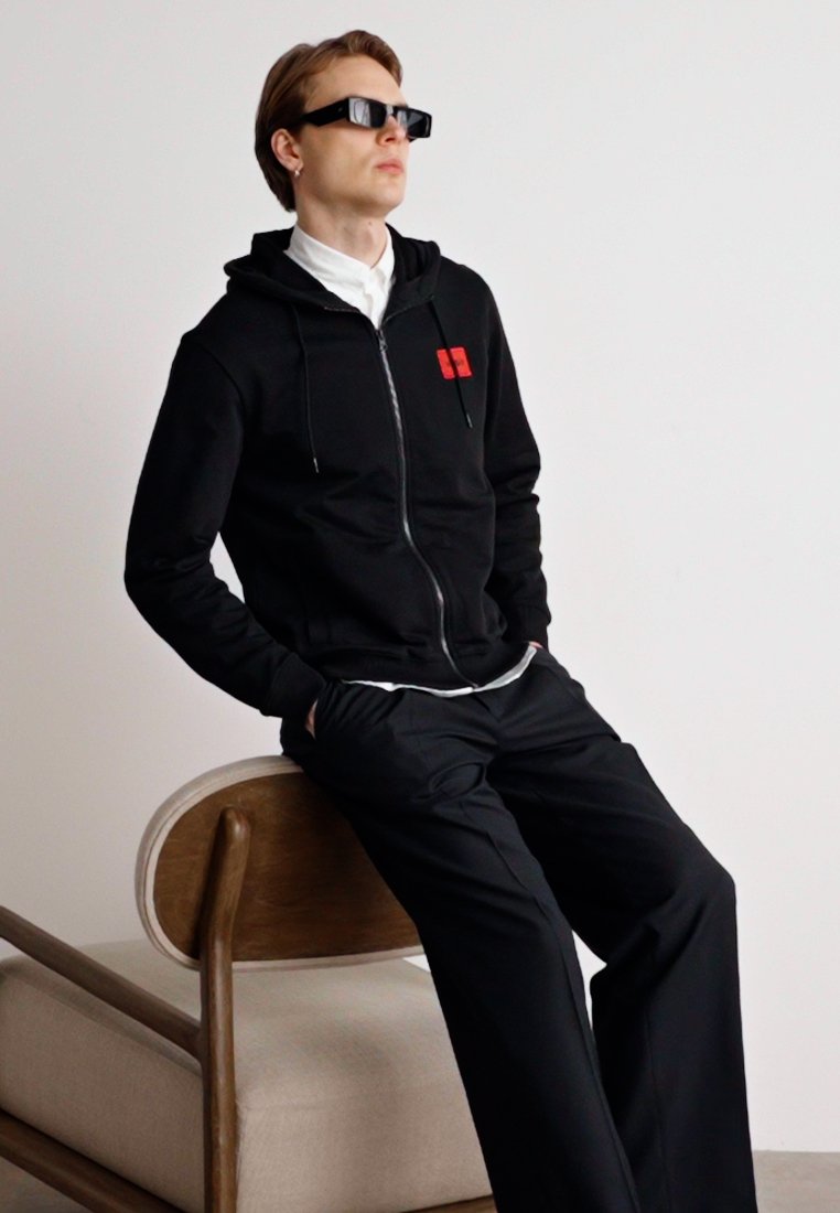 Veste à capuche noire zippée avec logo rouge, associée à un pantalon noir. Le modèle porte des lunettes de soleil noires et est assis sur une chaise en bois.