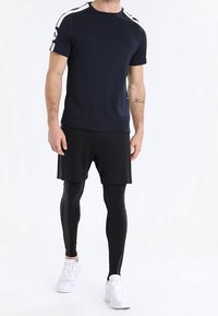 Marineblaues Sport-T-Shirt mit weißen Streifen auf den Schultern, kombiniert mit schwarzen Shorts und Leggings. Weiße Turnschuhe vervollständigen den Look.