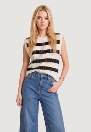 Femme aux longs cheveux blonds portant un top en maille crème sans manches avec des rayures horizontales noires et un jean bleu taille haute à jambes larges, posant avec la main dans la poche.