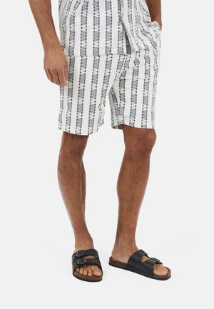 Uomo che indossa sandali neri con fibbia e pantaloncini bianchi con striscia verticale nera e motivo geometrico, in piedi con una mano in tasca.