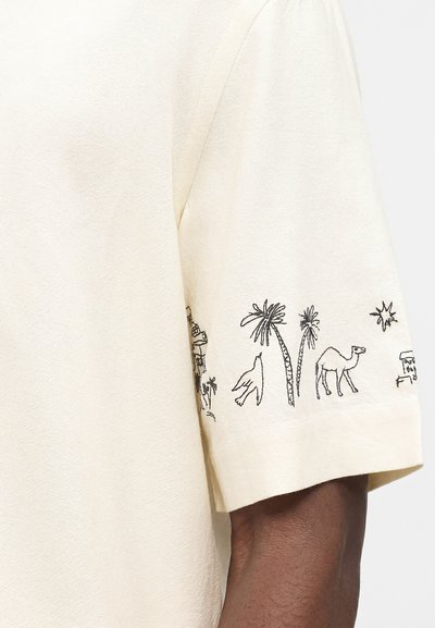 Κρεμ πουκάμισο με κοντά μανίκια και μαύρα σχεδιάκια γραμμών με φοινικόδεντρα, καμήλα, πουλί και μικρά κτίρια κατά μήκος της άκρης του μανικιού.