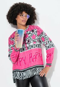 Kleurrijk shirt met gedurfde roze, zwarte en witte patronen, knopen en de tekst "Happy Happy." Gecombineerd met een glanzend, textured beker en zonnebril.