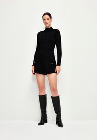 Schwarzes geripptes Langarmoberteil mit hohem Kragen, kombiniert mit maßgeschneiderten schwarzen Shorts mit zwei seitlichen Taschen. Schwarze kniehohe Stiefel mit Absatz.