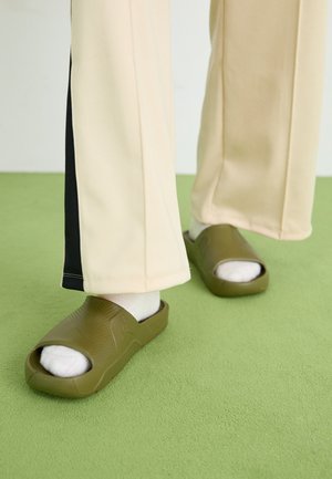 Des sandales en caoutchouc vertes avec un bout ouvert, une tige texturée et un motif décoratif, assorties à des chaussettes blanches à la cheville et un pantalon beige.