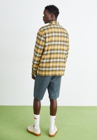 Camicia a quadri giallo e grigia, in cotone, maniche lunghe, abbinata a pantaloni corti grigi, calzini bianchi e sneakers beige su un pavimento verde.