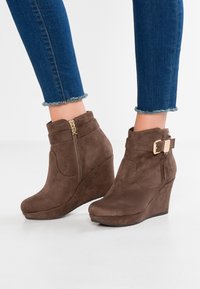 Stivali a zeppa in suede marrone con cerniera laterale e dettagli in metallo dorato. Caratterizzati da un risvolto con una fibbia decorativa. Superficie solida e testurizzata.
