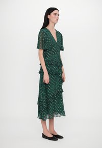 Groene midi-jurk met dierenprint, korte vleugelmouwen, een diepe V-hals, gelaagde ruches en een vloeiende, lichte stof. Zwarte platte schoenen.