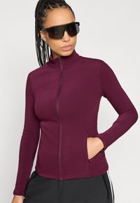 Μπουφάν μπορντό με φερμουάρ και μακριά μανίκια, κατασκευασμένο από απαλό ύφασμα. Διαθέτει ψηλό γιακά και πλαϊνές τσέπες, φορεμένο με σκουρόχρωμα γυαλιά ηλίου.