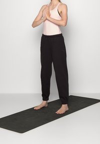 Czarne spodnie dresowe z elastycznymi mankietami i subtelnym logo, w zestawieniu z jasnoróżowym topem na ramiączkach. Figura stoi boso na ciemnej macie do jogi.