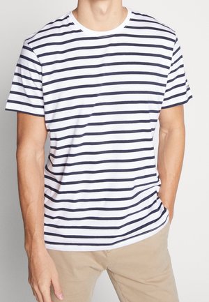 Man wearing a white short-sleeve T-shirt with black horizontal stripes and beige pants, one hand in pocket against a plain background.