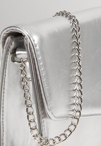 Sac à main en cuir argenté avec rabat et anse chaîne argentée attachée, présenté en gros plan.