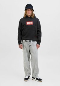 Giovane uomo con capelli lunghi, che indossa una felpa nera UFC, un cappello a secchiello, jeans larghi chiari e sneaker bianche e nere, in piedi contro uno sfondo chiaro uniforme.