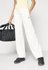 Λευκή φόρμα με ελαστικό κορδόνι, πλαϊνές τσέπες και μικρό λογότυπο στον μηρό, φορεμένη με εφαρμοστή ανοιχτόκίτρινη μπλούζα και μαύρα παπούτσια.