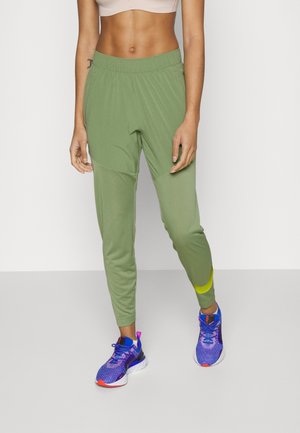 Femme portant un pantalon de jogging athlétique vert clair et des chaussures de course bleues, debout sur un fond blanc uni.