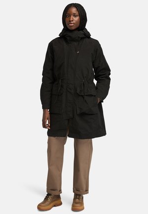 Black hooded parka with drawstring waist, featuring two front pockets, a sturdy fabric texture, and a knee-length design. Worn with tan trousers and boots.