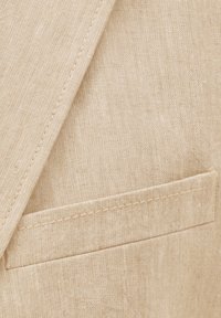 Beige linen jacket detail featuring a visible pocket and subtle texture, with prominent stitching and a clean, classic design.