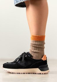 Zapatilla deportiva negra con texturas de gamuza y malla, suela rosa claro, cordones negros y talón acentuado en naranja, combinada con calcetines beige con canalé.
