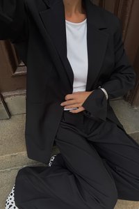 Črn blejzer preko bele srajce, v kombinaciji s črnimi hlačami. Oznake vključujejo dolge, široke rokave in delno vidne vzorčaste čevlje.