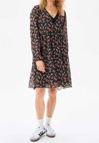 Nainen, joka pukeutuu mustaan kukalliseen polvipituiseen mekkoon, jossa on pitkät hihat, sekä valkoisiin tennareihin, joissa on mustat raidat, ja valkoisiin sukkiin.