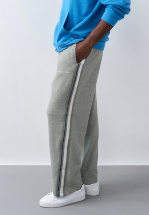 Άτομο με γκρι φόρμα Calvin Klein με ρίγες στο πλάι, μπλε φούτερ με κουκούλα, λευκά αθλητικά παπούτσια, στέκεται με το ένα χέρι στην τσέπη σε απλό γκρι πάτωμα.