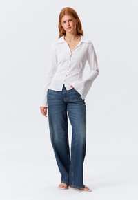 Camicia bianca con bottoni in tessuto strutturato, con colletto appuntito e maniche svasate, abbinata a jeans blu a gamba larga e sandali.