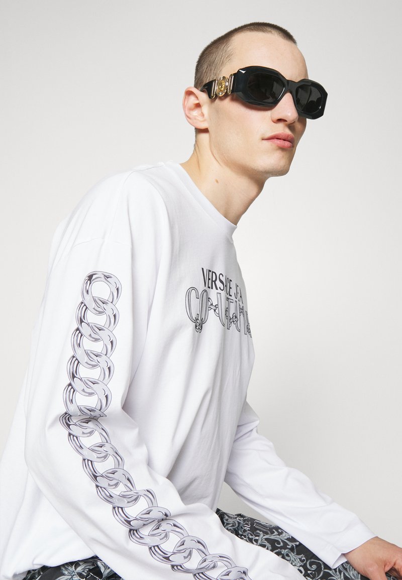 Camisa blanca de manga larga con gráficos de cadenas negras en las mangas y texto "VERSACE IS A COUTURE". El modelo lleva gafas de sol negras con acentos dorados.