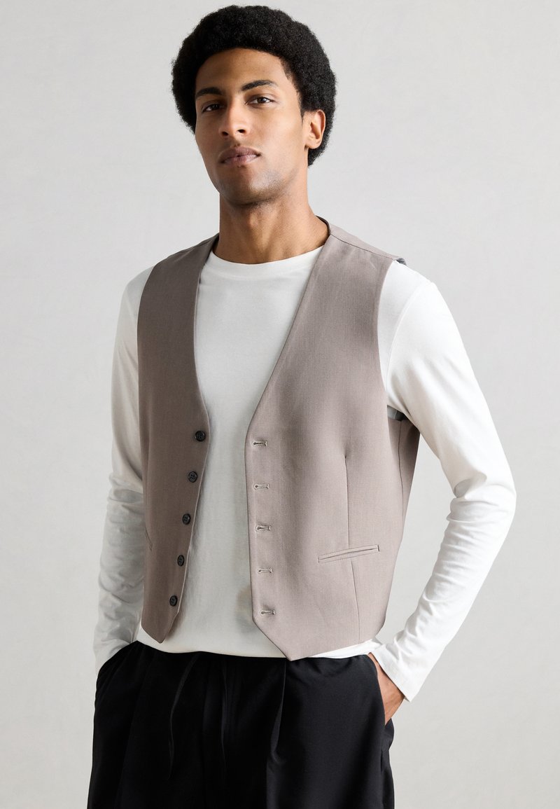 Γκρι υφασμάτινο γιλέκο με πέντε κουμπιά, φορεμένο πάνω από ένα λευκό πουκάμισο με μακρύ μανίκι. Διαθέτει κομψό σχέδιο και μία πλαϊνή τσέπη.