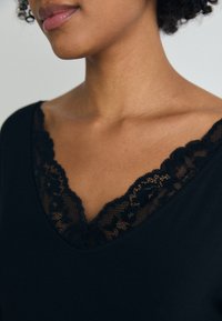 Svart topp med djup V-ringning och intrikat svart spetskant runt halsringningen. Den mjuka tygstrukturen framhäver den eleganta designen.