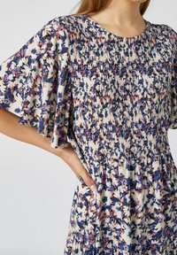 Robe fleurie avec un fond crème ornée de motifs violets et orange, corsage smocké et manches évasées. Tissu doux et coupe décontractée.