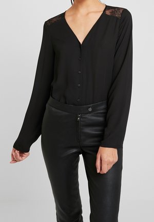 Frau trägt schwarze V-Ausschnitt-Bluse mit Spitzendetails an den Schultern und schwarze Lederhose mit Reißverschluss vorne und Knopfverschluss.