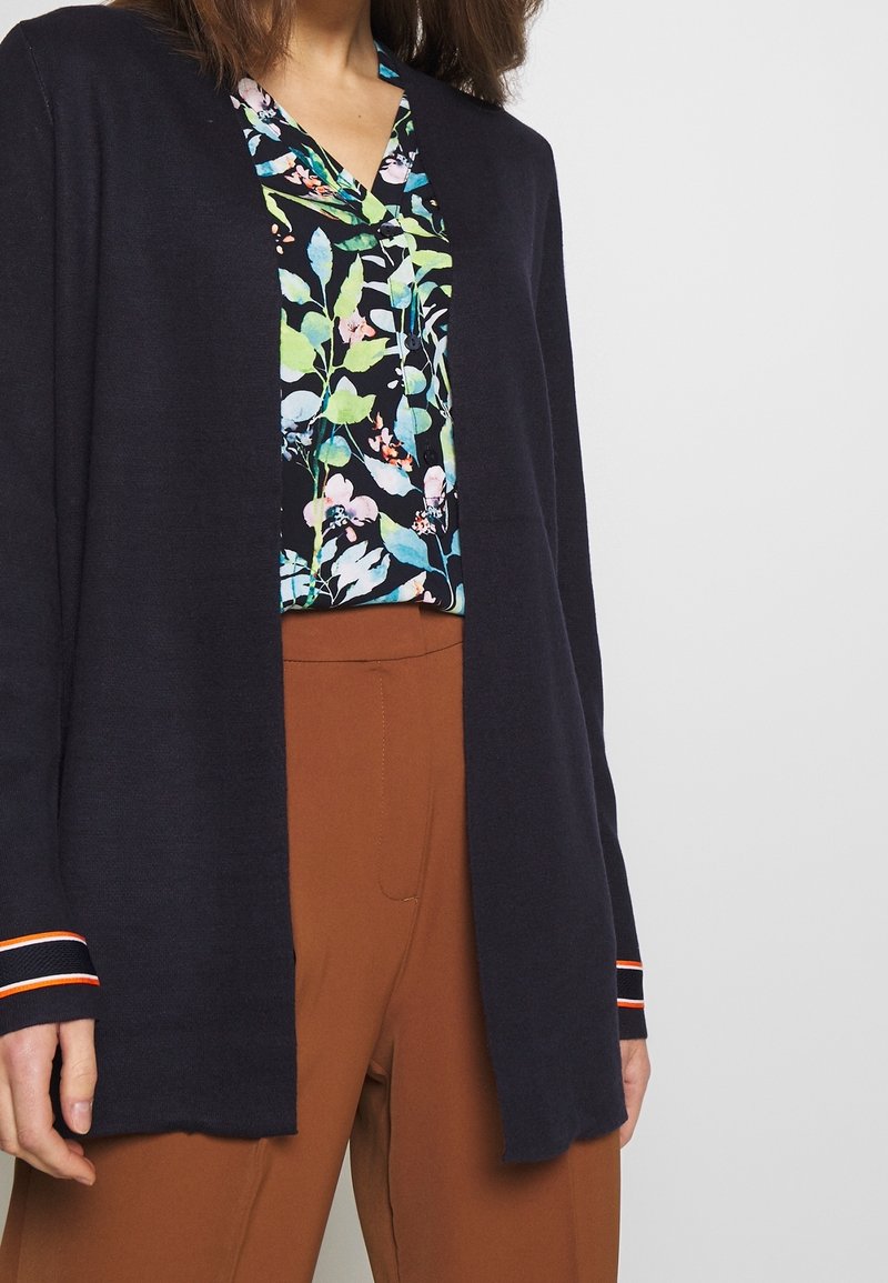 Marineblauer Cardigan mit offenem Frontbereich und langen Ärmeln, die gestreifte Akzente aufweisen, kombiniert mit einer blumigen Bluse und braunen High-Waist-Hosen.