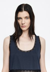 Top da donna blu scuro senza maniche con scollatura a barca e bordi in pizzo nero. Il tessuto è liscio, con un design a strati e delicati dettagli in pizzo.