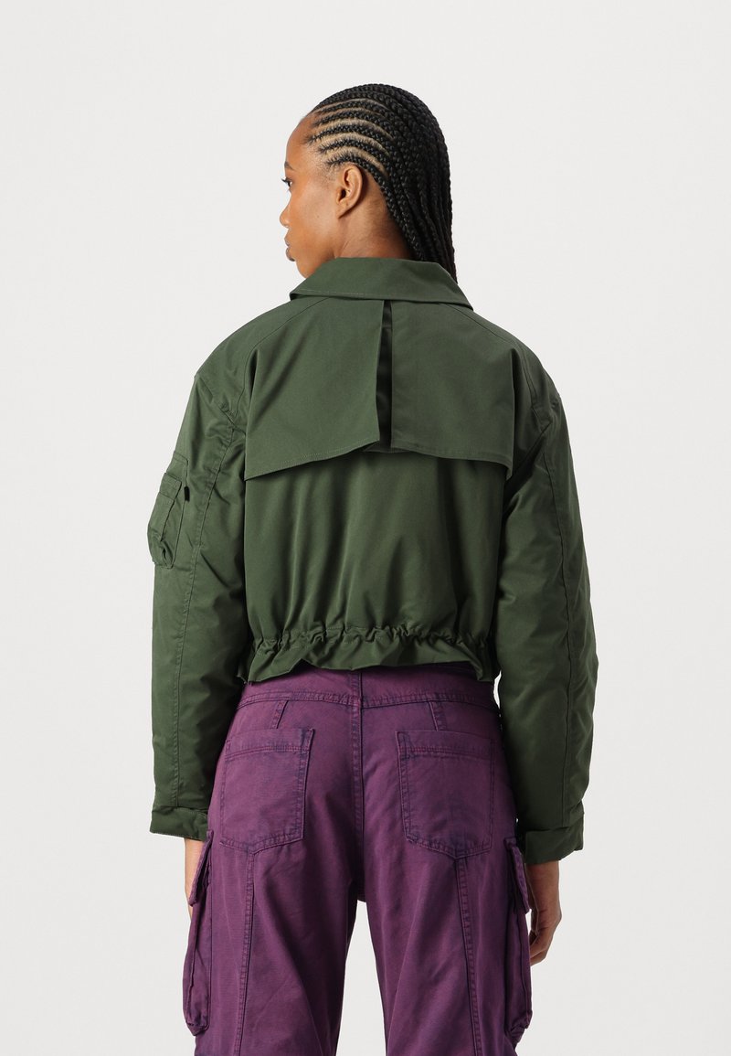 Chaqueta verde corta con una solapa en la parte posterior, cintura recogida y corte relajado, combinada con pantalones cargo morados que cuentan con bolsillos parche.