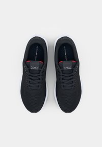 Baskets de sport noires avec une tige en mesh, des lacets plats et une semelle blanche. Présente un logo sur la languette et des accents rouges près du col.