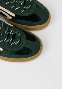 Sneaker verde con una texture in velluto e camoscio, caratterizzata da dettagli color crema, lacci testurizzati e una suola in gomma marrone con un design intrecciato.